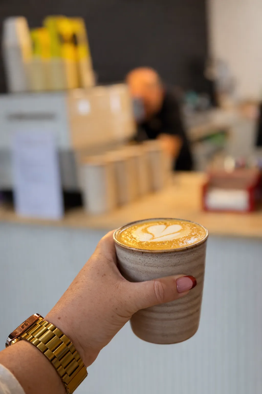 Hand holding a ceramic cup of coffee with heart latte art, barista behind counter